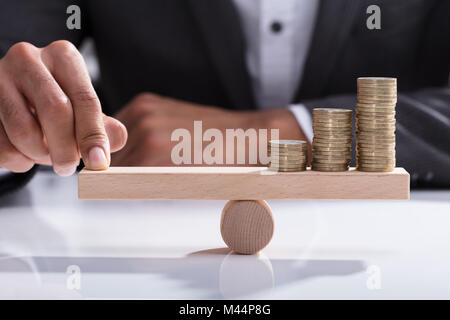 Close-up di un imprenditore la mano di bilanciamento delle monete impilate su altalena in legno con il dito sulla scrivania Foto Stock