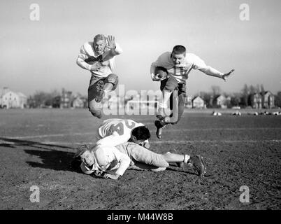 Due adolescenti giocatori di calcio senza caschi salto oltre i loro compagni di squadra replica di azione di gioco durante la pratica, ca. 1950. Foto Stock