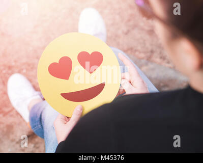 Cuore emoticon a portata di mano. Percosso la giovane donna tenendo la carta stampata o un cartone amore Smiley face. Buon San Valentino o anniversario del tema. Foto Stock