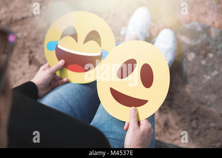 Donna che mantiene 2 emoticon di cartone in mano. Felice di ridere e di volti sorridenti. Due moderni di comunicazione e di espressione delle icone stampate su carta. Foto Stock