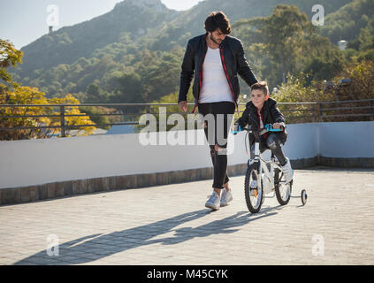 Giovane ragazzo in Bicicletta Equitazione con stabilizzatori, padre camminando accanto a lui Foto Stock