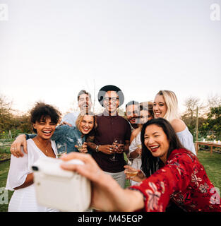 Gruppo di amici di scattare una foto con la fotocamera istantanea. Gli uomini e le donne al party in giardino prendendo un autoritratto. Foto Stock