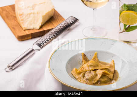 Ravioli di pasta con basilico e parmigiano sulla piastra. Parmigiano con una grattugia su un tagliere. Vino bianco e limonata Foto Stock