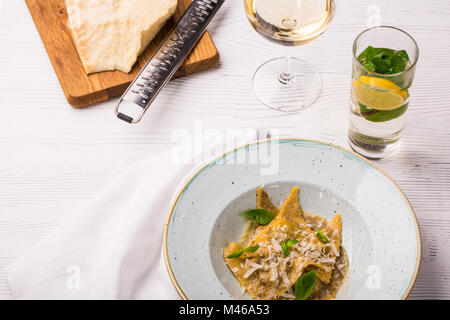 Ravioli di pasta con basilico e parmigiano sulla piastra. Parmigiano con una grattugia su un tagliere. Vino bianco e limonata Foto Stock