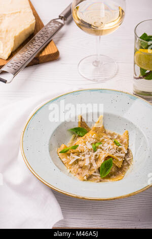 Ravioli di pasta con basilico e parmigiano sulla piastra. Parmigiano con una grattugia su un tagliere. Vino bianco e limonata. foto verticale Foto Stock