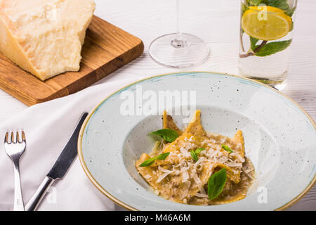 Ravioli di pasta con basilico e parmigiano sulla piastra. Parmigiano con una grattugia su un tagliere. Vino bianco e limonata Foto Stock