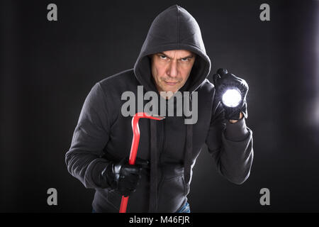 Ritratto di un ladro maschio tenendo la torcia elettrica e un palanchino su sfondo nero Foto Stock