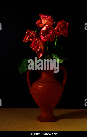 Cinque morendo Rose in un vaso di terracotta contro uno sfondo nero. Foto Stock