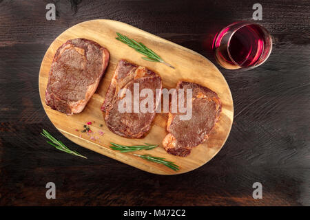 Una foto aerea di tre fette di carne cotta, filetti di manzo, ripresa dall'alto sul buio rustica con rametti di rosmarino, sale, pepe, vino, Foto Stock