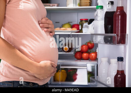 Close-up di donna incinta in piedi vicino al frigorifero pieno di cibi sani Foto Stock