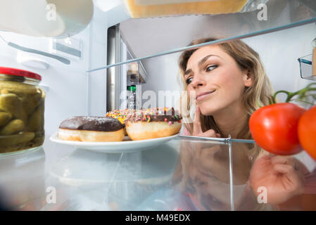 Close-up di un confuso giovane donna guardando le ciambelle in frigorifero aperto Foto Stock