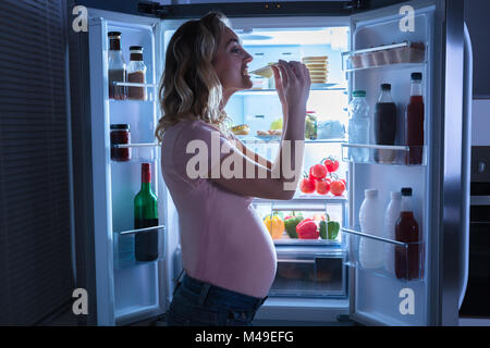 Close-up di una giovane donna incinta di mangiare fetta di formaggio nella parte anteriore del frigorifero Foto Stock