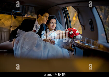 Ritratto di nozze. Felice giovane vestito elegantemente giovane è seduto in macchina e guardando attraverso la finestra. La sposa con il matrimonio bouquet di rose rosse e le gambe su lo sposo. Foto Stock