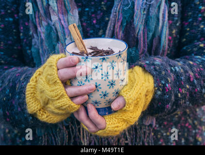 Womans mani con giallo guanti di lana tenendo una tazza di cioccolato caldo con un bastoncino di cannella e cioccolato fondente trucioli Foto Stock