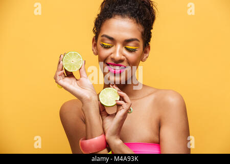 Ritratto di lieta afro american donna con il trucco alla moda tenendo due metà di lime fresco in entrambe le mani isolate su sfondo giallo Foto Stock