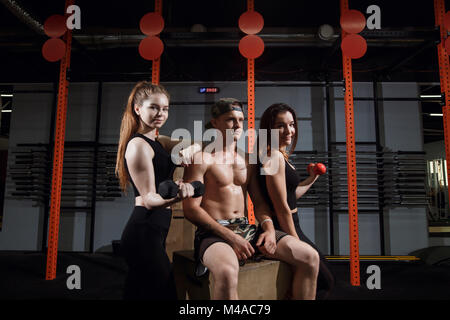 Gruppo di tre felice muscolare di femmine e maschi adulti seduti insieme sulla scatola come buoni amici in palestra con diffusore di grandi dimensioni in background dopo un difficile Foto Stock