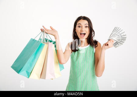 Ritratto di una ragazza sorpreso nel vestire holding shopping bags e mostrando mucchio di soldi banconote isolate su sfondo bianco Foto Stock