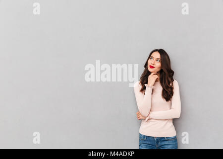 Ritratto orizzontale di affascinanti brunette femmina con lunghi capelli scuri puntella il mento con la mano e guardando a parte, su uno sfondo grigio spazio copia Foto Stock