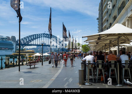 La gente camminare lungo e seduti al bar ristorante di Circular Quay Est con Harbour Bridge in distanza, Sydney, Nuovo Galles del Sud, Australia Foto Stock