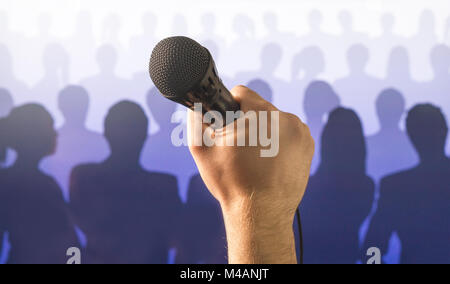 Parlare in pubblico e dare la parola concetto. Mano che tiene il microfono davanti a un pubblico di silhouette e la folla di gente. Il canto a microfono karaoke. Foto Stock