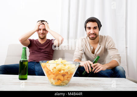 Due amici a guardare la TV con passione con birra e patatine Foto Stock