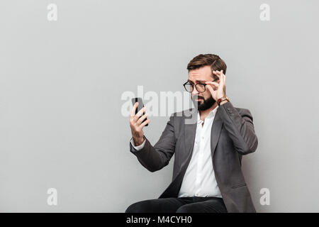 Ritratto orizzontale di imprenditore elegante guardando toccando lo smartphone occhiali mentre è seduto su una sedia in office isolate su uno sfondo grigio Foto Stock