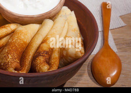 Tradizionale cibo oleoso in stile vintage: Frittelle con panna acida e un piatto di argilla con un cucchiaio di legno su un tavolo in legno di quercia Foto Stock