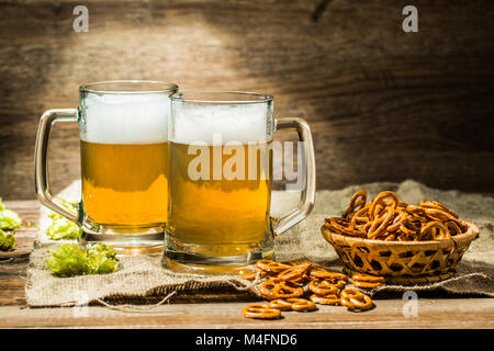 Due bicchieri di birra con la schiuma con hop e salatini su un tavolo di legno Foto Stock