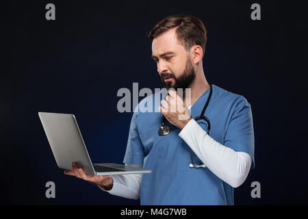 Ritratto di un concentrato medico maschio vestito in uniforme con uno stetoscopio guardando al computer portatile isolato su sfondo scuro Foto Stock