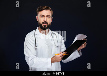 Ritratto di un maschio scioccato medico vestito in uniforme con uno stetoscopio holding notepad isolate su sfondo scuro Foto Stock