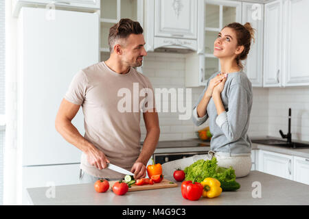 Ritratto di un sorridente amare giovane insalata di cottura insieme mentre in piedi su una cucina a casa, uomo taglio di vegetali Foto Stock
