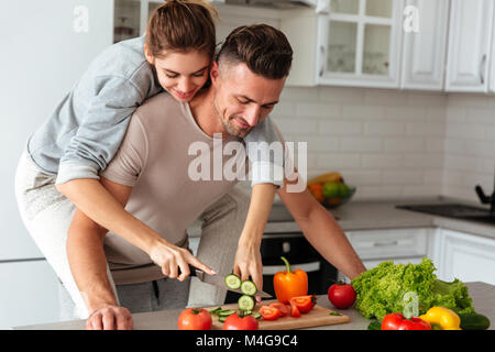 Ritratto di un sorridente amare giovane insalata di cottura insieme mentre in piedi su una cucina a casa, donna seduta sull uomo di spalle e vegetali di taglio Foto Stock