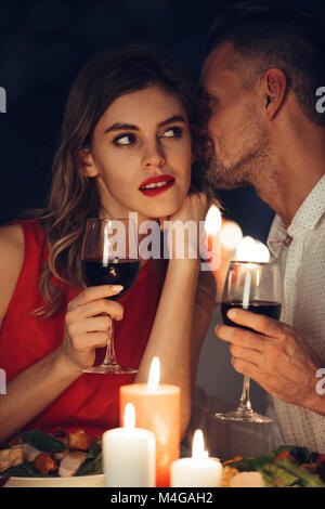 Curioso signora in abito rosso con bicchiere di vino ascoltando il suo uomo bello mentre hanno una romantica cena in serata Foto Stock