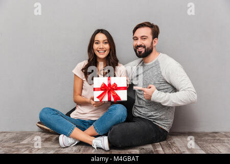 Ritratto di un sorridente coppia giovane tenendo presente la casella seduti insieme e puntare il dito su sfondo grigio Foto Stock