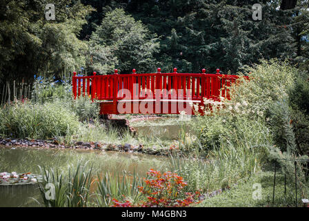 Un ponte rosso di oltre un flusso nei giardini botanici, Zagabria, Croazia. Il ponte è circondato da alberi e fiori Foto Stock