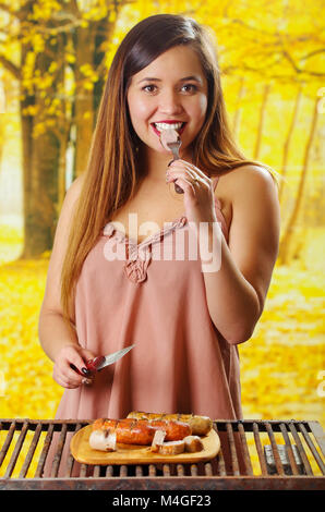 Close up di sorridere bella giovane donna di mangiare un pezzo di salsiccia con la forchetta e il coltello nelle sue mani, e salsicce alla griglia sul tagliere di legno, barbecue nel giardino. Salsicce bavaresi, in uno sfondo sfocato Foto Stock