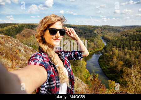 Giovane biondo sorridente, turista su una scogliera prendendo selfie immagine sullo sfondo del paesaggio autunnale con il fiume Berd. La Russia, Siberia, Salair Foto Stock