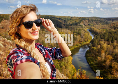 Giovane biondo sorridente, turista su una scogliera prendendo selfie immagine sullo sfondo del paesaggio autunnale con il fiume Berd. La Russia, Siberia, Salair Foto Stock