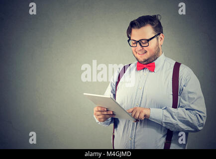 Giovane uomo cheeky in occhiali cercando felice durante la maschiatura tablet in posa su uno sfondo grigio. Foto Stock