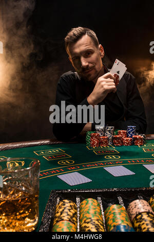 Bello il giocatore di poker con due assi nelle sue mani e patatine seduti al tavolo del poker in una stanza buia piena di fumo di sigaretta. Schede, trucioli, whisky, sigari Foto Stock