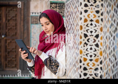 Donna musulmana lavorando su tablet in abbigliamento tradizionale con velo di colore rosso sulla sua testa accanto a un tradizionale arabesque parete marocchino Foto Stock