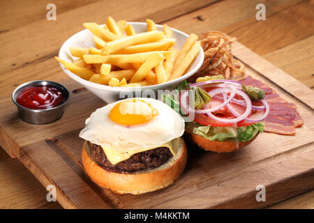 Burger con carne di manzo e uovo fritto sul vassoio in legno La piastra con patatine fritte sullo sfondo. Foto Stock