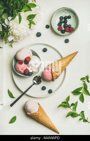 Flat-lay di rosa e fragola coconut ice cream scoop nella ciotola, dolce coni di cialda, peonia bouquet di fiori e di frutti di bosco freschi su sfondo bianco, t Foto Stock