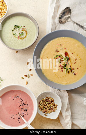 Flatlay con tre ciotole con colorati vegetariano zuppe cremose con broccoli, zucca e barbabietola guarnito con semi di zucca e olio d'oliva sul beige Foto Stock