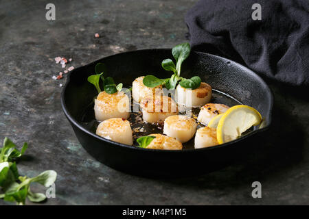 Capesante fritto con burro al limone salsa piccante in ghisa pan servito con insalata verde e del tessile igienico oltre il vecchio metallo scuro dello sfondo. Foto Stock