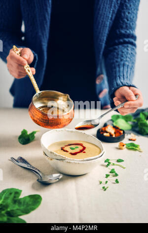 Una donna in blu cardigan drizzling una salsa sopra una ciotola di Bagno Turco, zuppa di lenticchie da una salsa di rame pan. Erbe, cucchiai e crostini accompagnare. Foto Stock