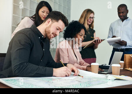 Giovane maschio architetto iscritto su blueprint ad una scrivania in un ufficio moderno con i colleghi di lavoro in background Foto Stock