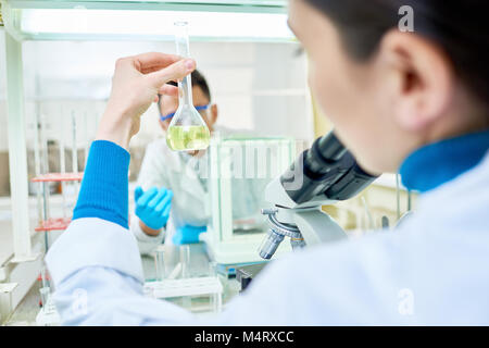 Sulla spalla vista di giovani dai capelli scuri azienda farmacia pallone con soluzione gialla in mano mentre è seduto alla scrivania, interno del laboratorio moderno sullo sfondo Foto Stock