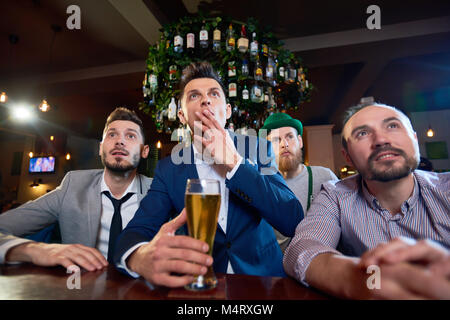 Il gruppo di amici di concentrato di guardare la partita di calcio mentre è seduto al banco bar e bere birra, uomo barbuto indossando il costume leprechaun in piedi Foto Stock