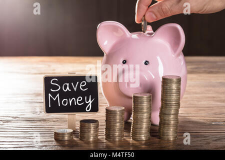 Persona inserimento di moneta in Piggybank con pile di monete sul tavolo di legno che mostra risparmiare denaro Concept Foto Stock